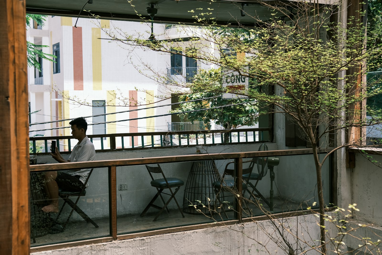 Ban công quán cafe Việt — người ngồi đọc điện thoại, cây xanh